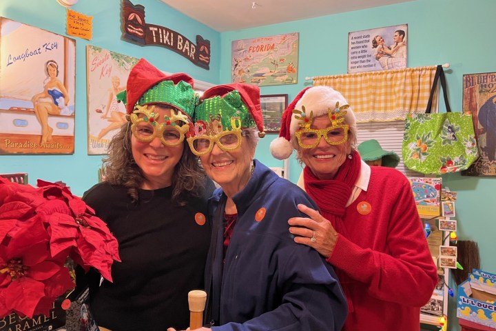 Three smiling women in festive glasses and holiday attire standing indoors.