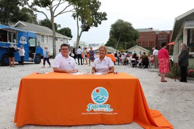 Two people at an orange table with 'Discover Sarasota Tours' at a community event.
