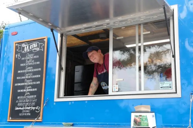 Person in blue food truck serving, with menu board beside them.