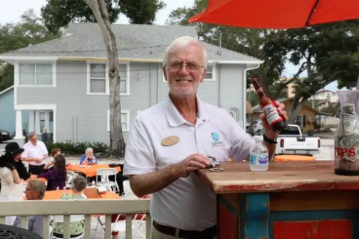 Man at outdoor party, holding a beer bottle behind a colorful bar.