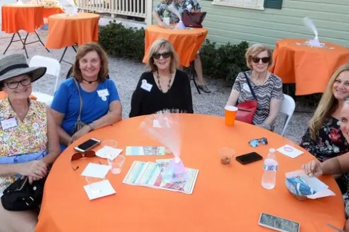 Six women seated at a round table with orange tablecloths outside.