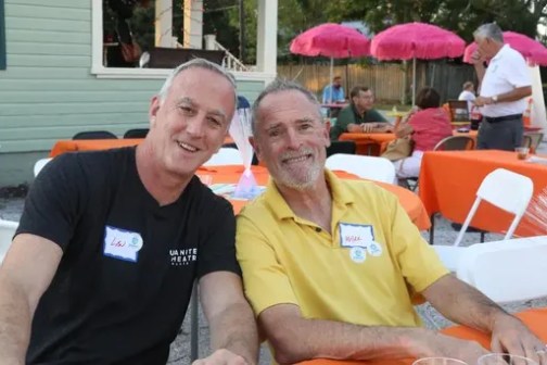 Two men smiling at an outdoor event with tables and umbrellas.