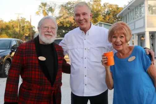 Three smiling people posing outdoors, one holding an orange cup, with trees and buildings in the background.