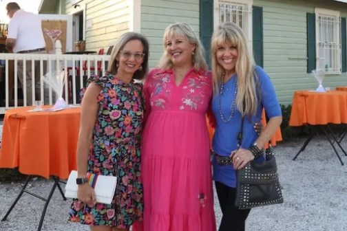 Three women posing outside near a house with orange tablecloths on tables.