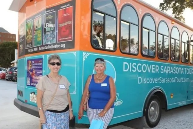 Two women smiling in front of a colorful tour bus in a parking lot.