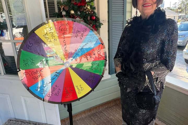 Woman in festive attire stands next to a colorful prize wheel on a porch.
