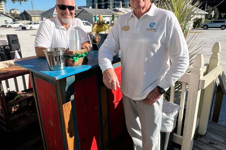 Two men in white shirts stand by a colorful outdoor bar on a sunny day.
