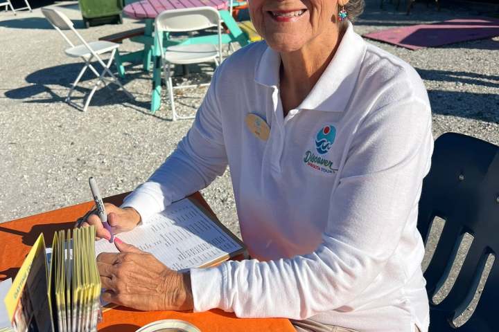 Smiling person wearing sunglasses at outdoor table with brochures and name tags.