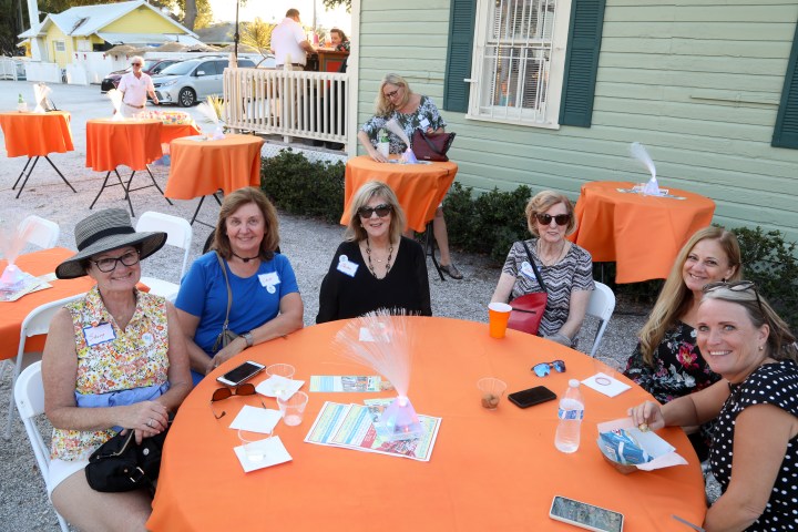 Six women seated at a table with orange tablecloths outdoors.