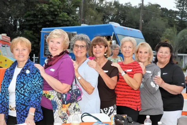 Seven women smiling, standing in a row outdoors near food trucks.