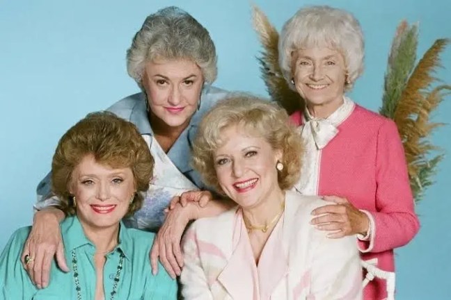 Four older women smiling, posing closely in colorful outfits.