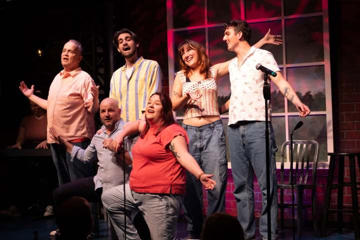 Group of six people performing on stage, smiling, with microphones.