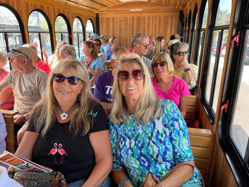 Group of people sitting in a wooden tram, two women in sunglasses smiling in the foreground.