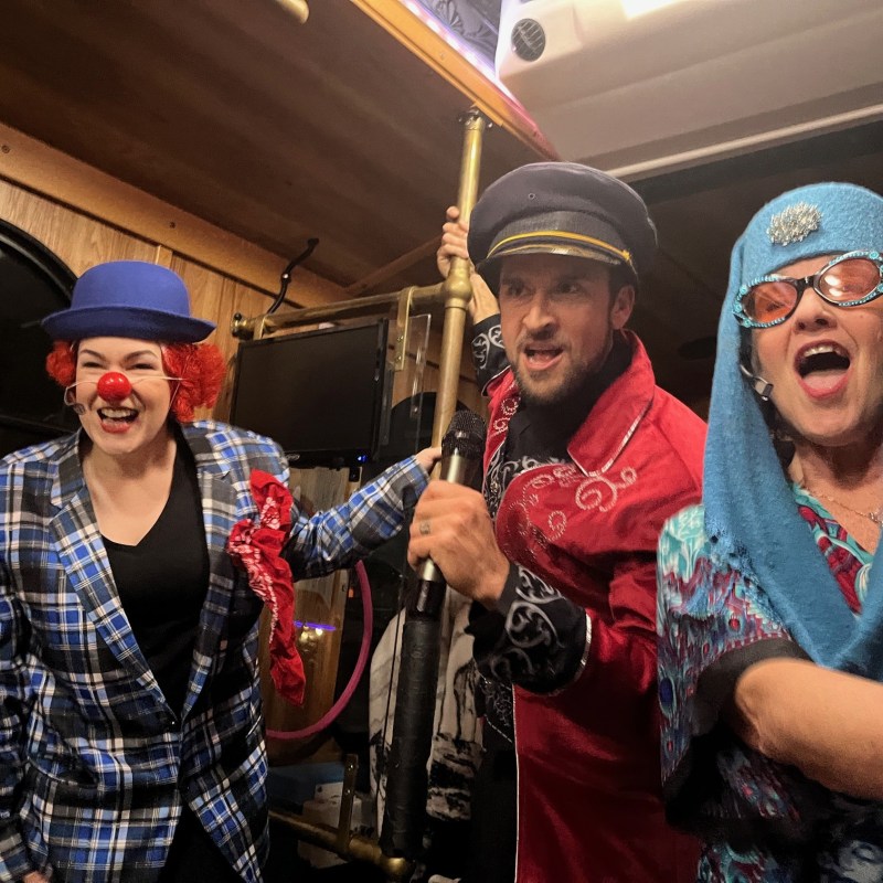 Three people in colorful costumes, including a clown, inside a vehicle, smiling and posing energetically.