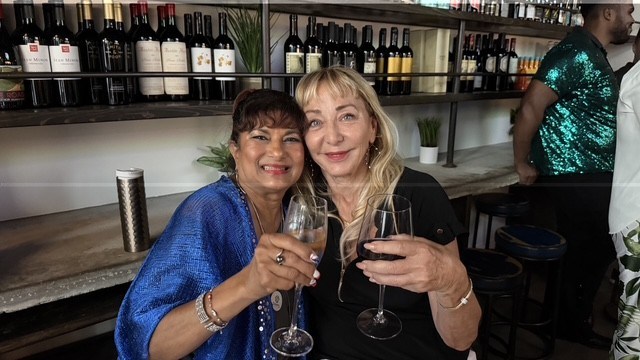 Two women smiling and holding wine glasses in a bar with wine bottles in the background.