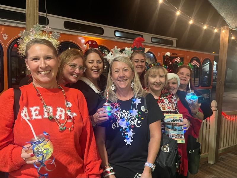 Group of women smiling, wearing festive attire, holding drinks, in front of an orange trolley at night.