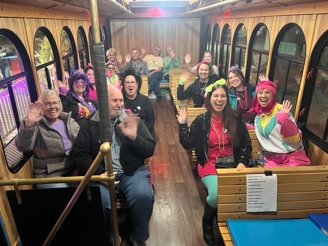 Group of smiling people in colorful outfits waving inside a bus.