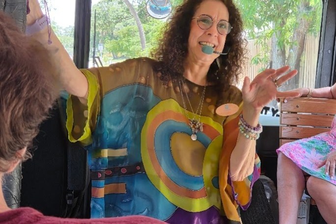 Woman in colorful outfit speaking on a bus, holding a microphone. A small white dog sits nearby.