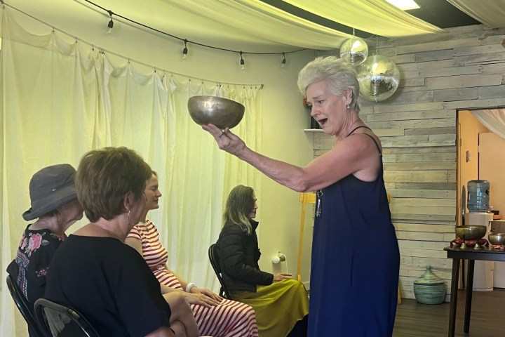 Woman using sound bowl in front of seated group in a decorated room.