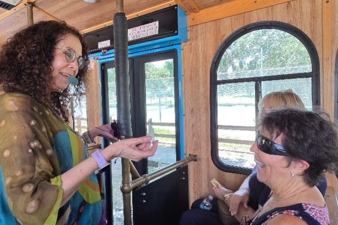 Woman in colorful shawl engaging with seated passengers on a wooden interior bus.