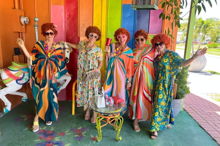 Five people in colorful dresses and wigs pose joyfully against a vibrant wall.