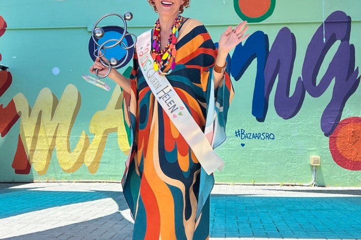 Person in colorful dress and sash with trophy, standing under blue canopy.