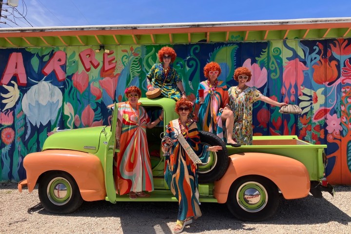 Five people with red wigs and colorful outfits on a vintage truck in front of a vibrant mural.
